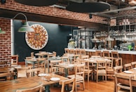 A rustic restaurant interior with wooden tables and chairs, and a brick wall. The dining area has table settings with plates and cutlery, and a bar area with various bottles and hanging wine glasses. On the wall, there's a colorful mosaic or painting of a sun face. The lighting is provided by green hanging lamps.