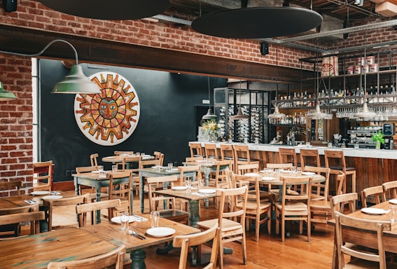 A rustic restaurant interior with wooden tables and chairs, and a brick wall. The dining area has table settings with plates and cutlery, and a bar area with various bottles and hanging wine glasses. On the wall, there's a colorful mosaic or painting of a sun face. The lighting is provided by green hanging lamps.