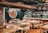 A rustic restaurant interior with wooden tables and chairs, and a brick wall. The dining area has table settings with plates and cutlery, and a bar area with various bottles and hanging wine glasses. On the wall, there's a colorful mosaic or painting of a sun face. The lighting is provided by green hanging lamps.