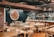 A rustic restaurant interior with wooden tables and chairs, and a brick wall. The dining area has table settings with plates and cutlery, and a bar area with various bottles and hanging wine glasses. On the wall, there's a colorful mosaic or painting of a sun face. The lighting is provided by green hanging lamps.