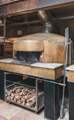 A rustic wood-fired oven with a mosaic tiled exterior is situated in a commercial kitchen setting. The oven has a metallic vent pipe extending from the top. Below the oven, there is a storage area filled with firewood logs. A long pizza peel rests against the oven.