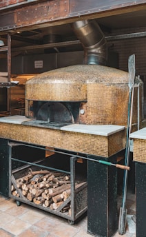 A rustic wood-fired oven with a mosaic tiled exterior is situated in a commercial kitchen setting. The oven has a metallic vent pipe extending from the top. Below the oven, there is a storage area filled with firewood logs. A long pizza peel rests against the oven.