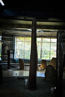 The image depicts an indoor exhibition space featuring several tall, upright stone monuments. These stones are displayed on a gravel-covered floor and are arranged with small numbered signs in front of them. There is a large window in the background allowing natural light to filter in, illuminating the stones and surroundings. The ceiling has an industrial look, and the overall atmosphere is that of a museum or gallery.