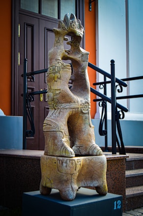 An intricately carved abstract sculpture made of stone or ceramic stands on a dark pedestal. It features organic shapes and patterns, resembling intertwined figures or animals. The sculpture is outside, in front of a doorway and next to stairs with black railings.