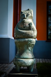 A large metallic sculpture resembling a stylized human or fantasy creature stands outdoors against a backdrop of a building with white, orange, and grey walls. The sculpture has intricate carvings and patterns, with arms folded and a stoic expression on its face. The sculpture is placed on a paved walkway next to some grass.