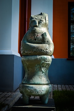 A large metallic sculpture resembling a stylized human or fantasy creature stands outdoors against a backdrop of a building with white, orange, and grey walls. The sculpture has intricate carvings and patterns, with arms folded and a stoic expression on its face. The sculpture is placed on a paved walkway next to some grass.