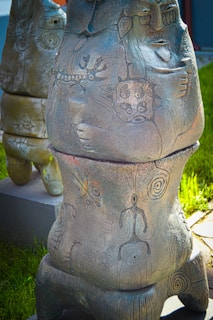 A sculpture with intricate carvings displaying abstract and tribal art. The design includes various symbols and patterns, such as spirals and figures, giving an ancient or mystical feel. The sculpture stands in an outdoor setting on a grassy background.