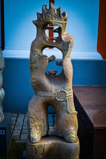 An intricately carved stone sculpture with a totem-like design. It features abstract shapes and symbols, with a prominent open space in the center. The surface is textured with various patterns and motifs, suggesting cultural or artistic significance. The sculpture stands on a block base and is surrounded by outdoor elements.
