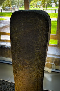 A large stone slab standing vertically, covered with inscriptions or carvings that resemble ancient runes. The stone is displayed indoors with large windows behind it, providing natural lighting. Outside, green grass and trees are visible, giving a sense of a museum or historical display area.