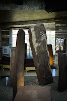 Large standing stones are placed indoors, surrounded by small gravel. The stones exhibit rough surfaces with varied textures and colors, prominently featuring a stone with a large white patch. Behind, modern furniture such as sofas is visible, set against a backdrop of windows letting in natural light.