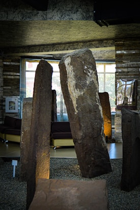 Large standing stones are placed indoors, surrounded by small gravel. The stones exhibit rough surfaces with varied textures and colors, prominently featuring a stone with a large white patch. Behind, modern furniture such as sofas is visible, set against a backdrop of windows letting in natural light.