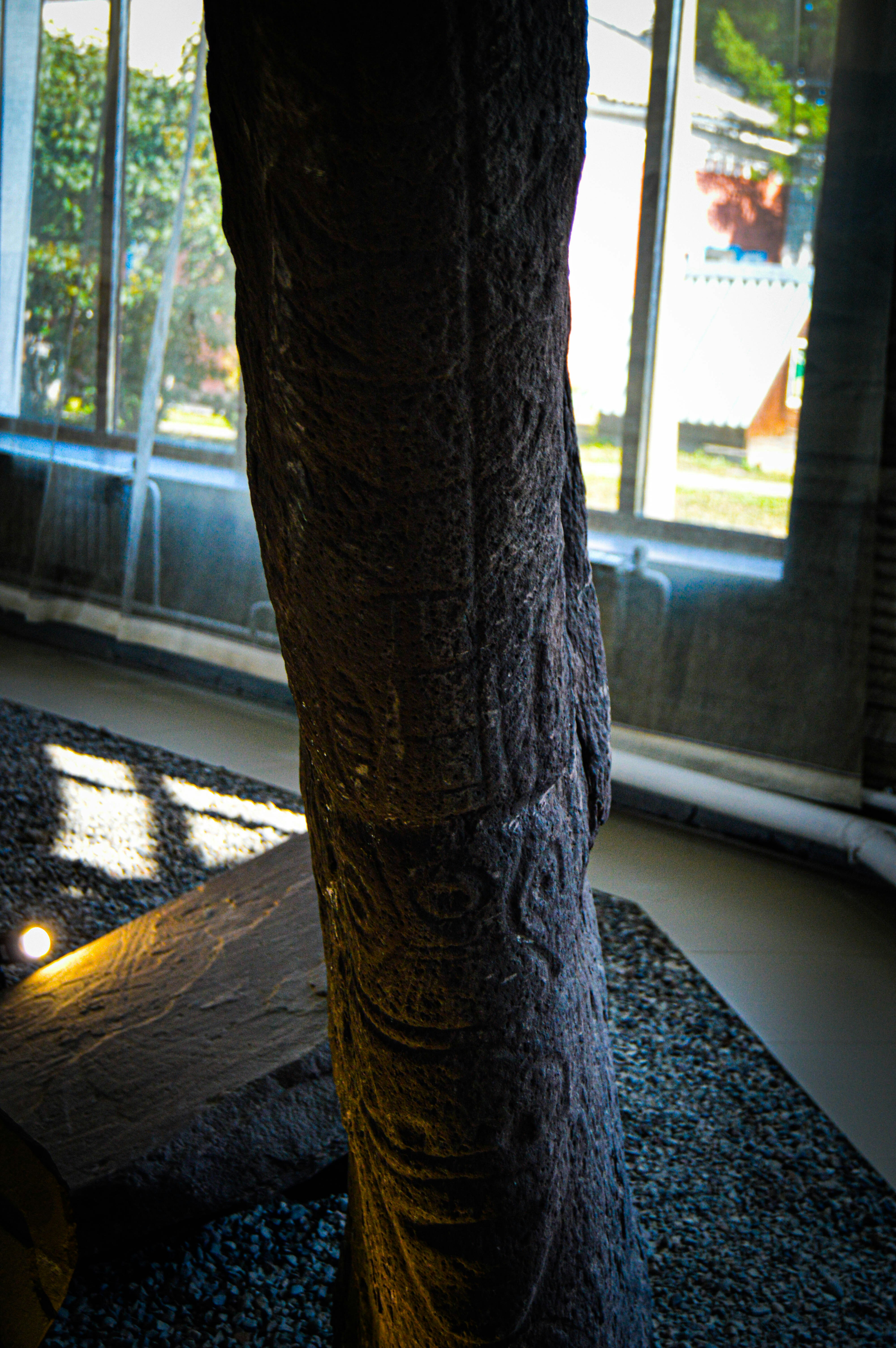 A large, textured stone pillar is positioned indoors against a backdrop of large windows allowing natural light to filter in. The surface of the pillar is sculpted with intricate patterns, and it rests on a gravel bed. Some light fixtures are visible, illuminating the stone from below.