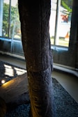 A large, textured stone pillar is positioned indoors against a backdrop of large windows allowing natural light to filter in. The surface of the pillar is sculpted with intricate patterns, and it rests on a gravel bed. Some light fixtures are visible, illuminating the stone from below.