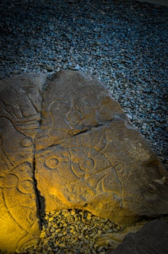 A weathered stone slab featuring intricate carvings rests on a bed of pebbles. The carvings depict abstract shapes and figures, illuminated by a soft, warm light that creates a contrast with the cool, bluish hue of the surrounding pebble surface.