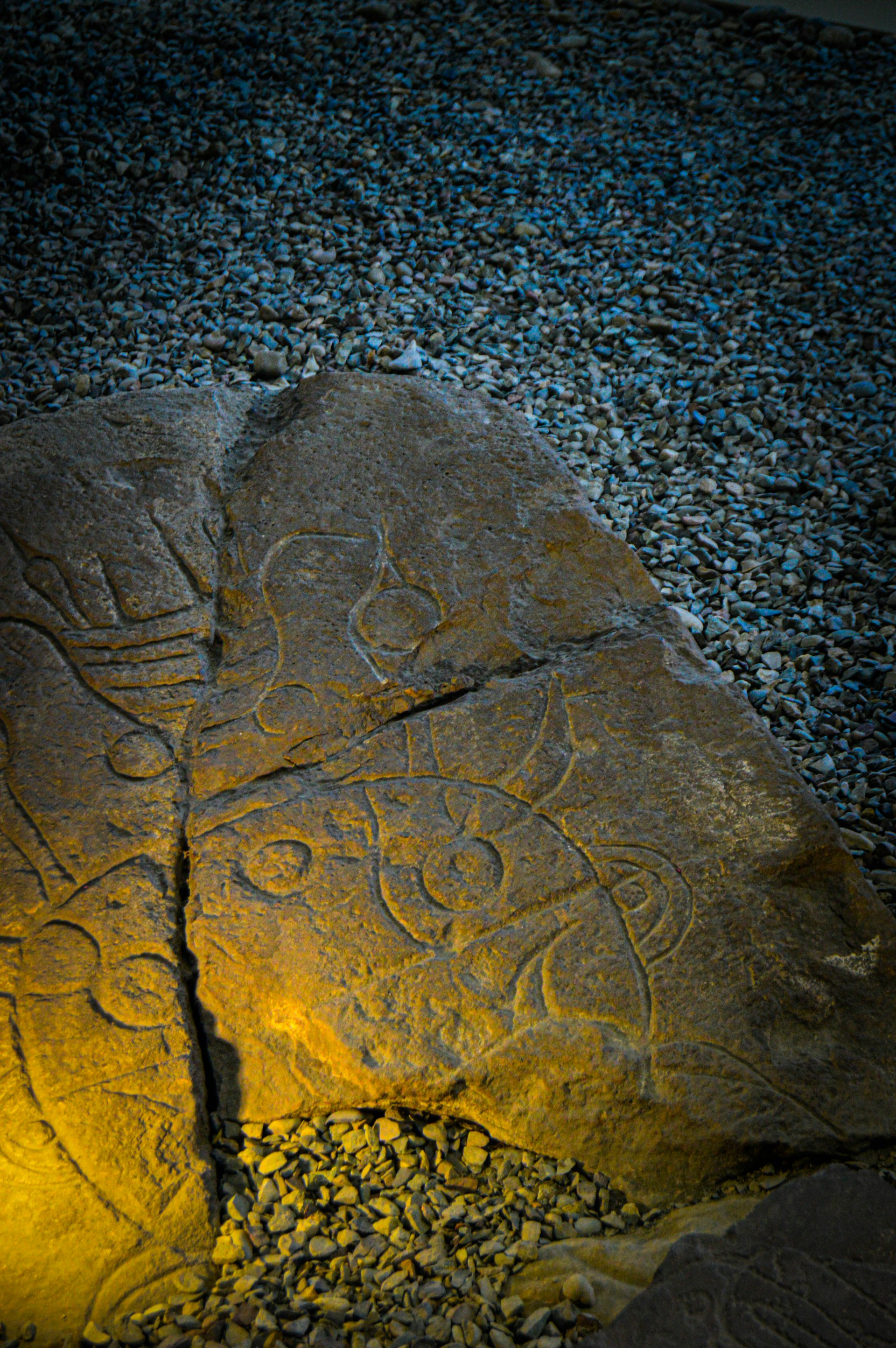 A weathered stone slab featuring intricate carvings rests on a bed of pebbles. The carvings depict abstract shapes and figures, illuminated by a soft, warm light that creates a contrast with the cool, bluish hue of the surrounding pebble surface.