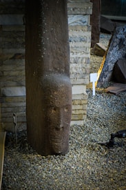 A stone figure resembling a stylized human face, possibly part of a sculpture, is positioned vertically on a gravel-covered surface. It is surrounded by a variety of stone and brick textures, including stacked stone blocks behind it. The lighting creates a contrast between the dark brown hue of the stone figure and the lighter background elements.
