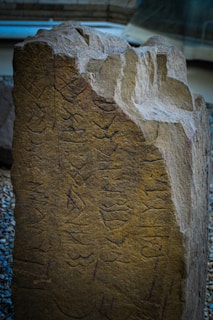 A large stone slab with ancient carvings or inscriptions on its surface, possibly depicting runes or symbols. The texture of the stone is rough, with a natural, weathered appearance. Surrounding the stone are small pebbles, and the background is slightly blurred, suggesting an indoor setting with architectural elements.