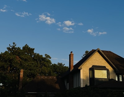 A house with a sloped roof and chimney is partially shaded and set against a backdrop of trees under a clear blue sky. Soft sunlight illuminates part of the facade, highlighting the stonework and window details.
