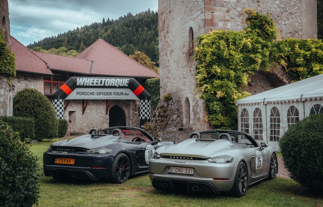 Two Porsche sports cars are parked on a grassy area in front of an old stone building with ivy. A 'Wheeltorque Porsche Spyder Tour' banner is displayed above the entrance, creating a blend of historical architecture and modern automobile design.
