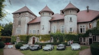 A historic, ivy-covered stone chateau with multiple turrets stands as the backdrop. In front, there is a neatly trimmed lawn displaying five luxury sports cars parked side by side. A group of people is gathered near the cars, possibly engaged in conversation. The atmosphere is serene and elegant, with overcast skies above.