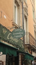 An outdoor view of a traditional Italian building showcasing its old architectural style. There's a sign for 'Osteria Locandina Cappello' hanging above, with green awnings beneath it. The building has warm-toned walls, ornate windows, and decorative plants.
