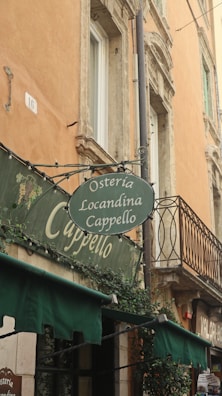 An outdoor view of a traditional Italian building showcasing its old architectural style. There's a sign for 'Osteria Locandina Cappello' hanging above, with green awnings beneath it. The building has warm-toned walls, ornate windows, and decorative plants.