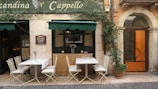 An outdoor seating area of a small restaurant or cafe with two tables, each set with tablecloths and glassware, surrounded by white chairs. Potted plants are visible, including one with a climbing vine next to an arched wooden door. The building facade is rustic with a sign that reads 'Osteria Locanda Cappello' above the tables under a green awning.