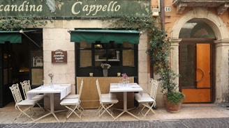 An outdoor seating area of a small restaurant or cafe with two tables, each set with tablecloths and glassware, surrounded by white chairs. Potted plants are visible, including one with a climbing vine next to an arched wooden door. The building facade is rustic with a sign that reads 'Osteria Locanda Cappello' above the tables under a green awning.
