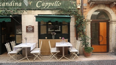 An outdoor seating area of a small restaurant or cafe with two tables, each set with tablecloths and glassware, surrounded by white chairs. Potted plants are visible, including one with a climbing vine next to an arched wooden door. The building facade is rustic with a sign that reads 'Osteria Locanda Cappello' above the tables under a green awning.