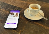 A smartphone displaying a dating app interface is placed on a wooden table next to a small cup of espresso on a saucer with a spoon.