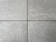 Four square cement tiles arranged in a grid pattern with visible seams between them. The surface of each tile has a textured, speckled appearance with shades of gray and subtle variations in tone.