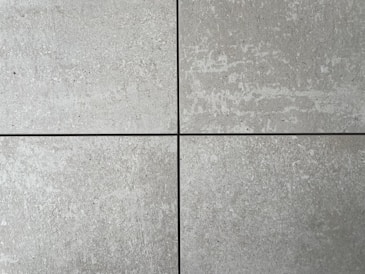 Four square cement tiles arranged in a grid pattern with visible seams between them. The surface of each tile has a textured, speckled appearance with shades of gray and subtle variations in tone.