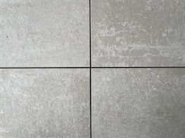 Four square cement tiles arranged in a grid pattern with visible seams between them. The surface of each tile has a textured, speckled appearance with shades of gray and subtle variations in tone.