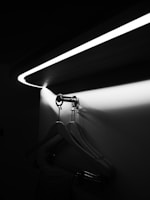 A black and white photograph shows a sleek, modern closet area with wooden hangers hanging on a metallic rod. Above, an elegantly curved LED light casts a soft glow and illuminates the space, creating contrasting shadows on the wall.