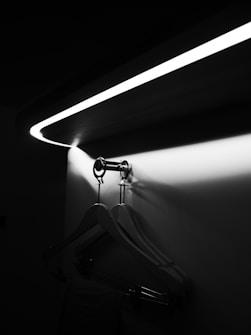 A black and white photograph shows a sleek, modern closet area with wooden hangers hanging on a metallic rod. Above, an elegantly curved LED light casts a soft glow and illuminates the space, creating contrasting shadows on the wall.