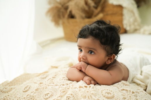 A baby with dark hair lies on a cream-colored crocheted blanket, gazing thoughtfully with fingers in their mouth. Soft, natural lighting enhances the cozy and serene atmosphere, complemented by the blurred background of earthy tones, likely from decor elements.