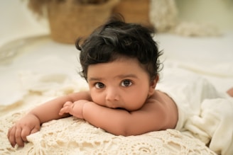 A baby with dark curly hair lies on its stomach on a cream-colored crocheted blanket. The baby has large expressive eyes and is looking directly at the camera. The background is a soft, neutral color with a wicker basket and plush items out of focus.
