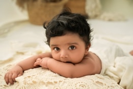 A baby with dark curly hair lies on its stomach on a cream-colored crocheted blanket. The baby has large expressive eyes and is looking directly at the camera. The background is a soft, neutral color with a wicker basket and plush items out of focus.