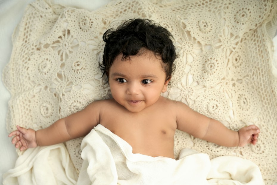 A smiling baby with dark hair lies on a cream-colored, intricately patterned crochet blanket. The baby is wrapped in a soft white blanket, spreading its arms outward.