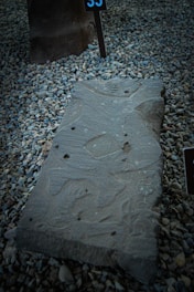 A carved stone slab is placed on a bed of small gravel rocks. The slab features intricate carvings and patterns that are weathered over time. Nearby, a numbered sign is positioned upright in the gravel.