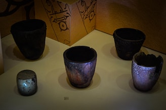 Several ancient pottery pieces are displayed on a flat surface with a beige textured background. The pottery varies in size and texture, with some surfaces appearing smooth and others having intricate patterns. There is also visible wall art featuring abstract designs in the background.