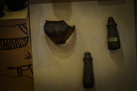 Three ancient artifacts are displayed on a beige surface. The left artifact resembles a broken dark bowl with intricate patterns. The two objects on the right appear to be stone tools or ceremonial items with a conical shape and a rough, textured surface.