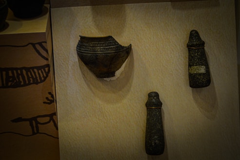 Three ancient artifacts are displayed on a beige surface. The left artifact resembles a broken dark bowl with intricate patterns. The two objects on the right appear to be stone tools or ceremonial items with a conical shape and a rough, textured surface.