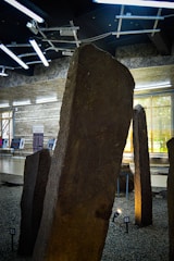 Several large stone monoliths are displayed indoors on a gravel surface. The ceiling features modern lighting fixtures, and informational boards are visible in the background, positioned near large windows that let in natural light.