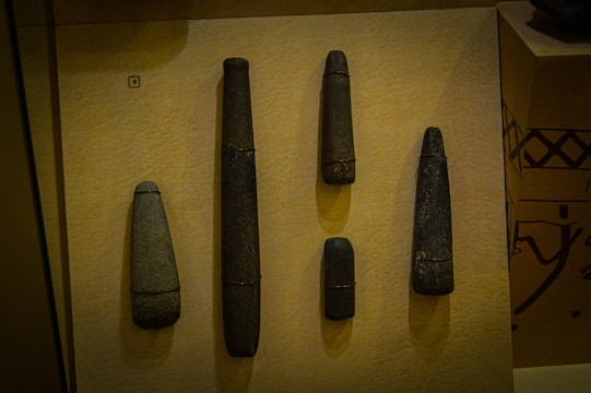 Five stone tools of varying sizes are displayed vertically against a textured tan background. Each tool is elongated and cylindrical, with some showing narrower tips. The stones appear dark and weathered, with subtle variations in color and texture.