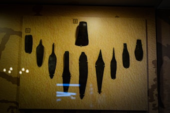 An exhibit featuring an array of ancient tools displayed against a light yellow background. The tools vary in shape and size, arranged in a symmetrical pattern, and appear to be made of dark metal. Each tool is individually mounted, and small numbered labels are placed above them.