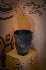 A dark, textured ceramic vase with cracks, sitting on a decorated pedestal against a background with abstract, tribal-like patterns and earthy tones.