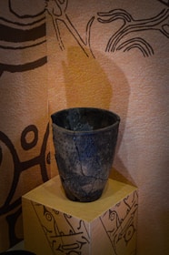 A dark, textured ceramic vase with cracks, sitting on a decorated pedestal against a background with abstract, tribal-like patterns and earthy tones.