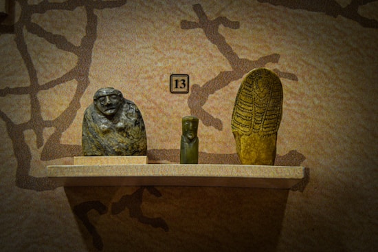 Three ancient artifacts are displayed on a wooden shelf against a textured wall. The items include a sculpted stone head with an abstract face, a small green figurine in the center, and an elongated stone object with intricate designs resembling a stylized figure or mummy.