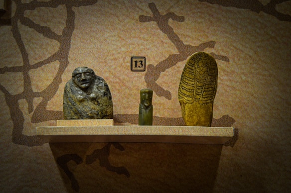 Three ancient artifacts are displayed on a wooden shelf against a textured wall. The items include a sculpted stone head with an abstract face, a small green figurine in the center, and an elongated stone object with intricate designs resembling a stylized figure or mummy.